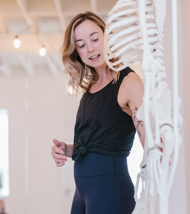 Alice, is examining a human skeleton model in a bright studio space. Her attention is on the skeletal details, possibly during an anatomy class for yoga, highlighting the educational aspect of her teaching.