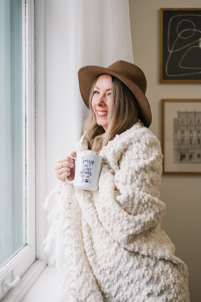 Sammy in a hat wrapped in a white blanket holding a mug and looking out the window