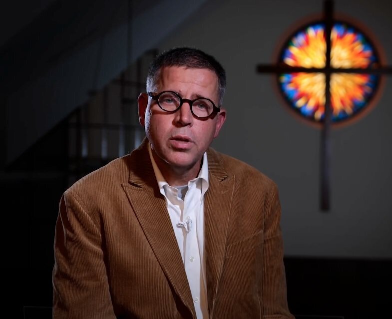 A person in a suit and glasses being interviewed 