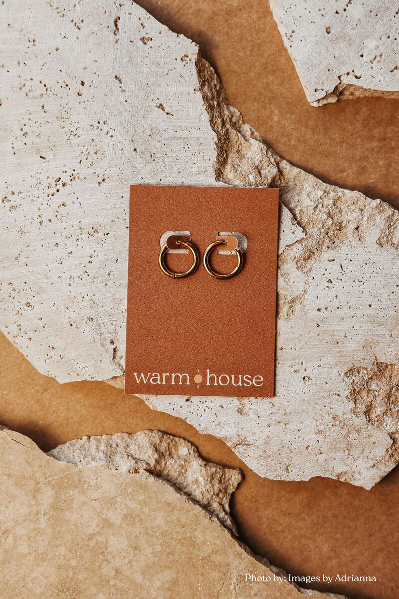 A custom branding earring card for the brand "warm House" holds two gold hoop earrings and sits atop a chiseled sandstone surface