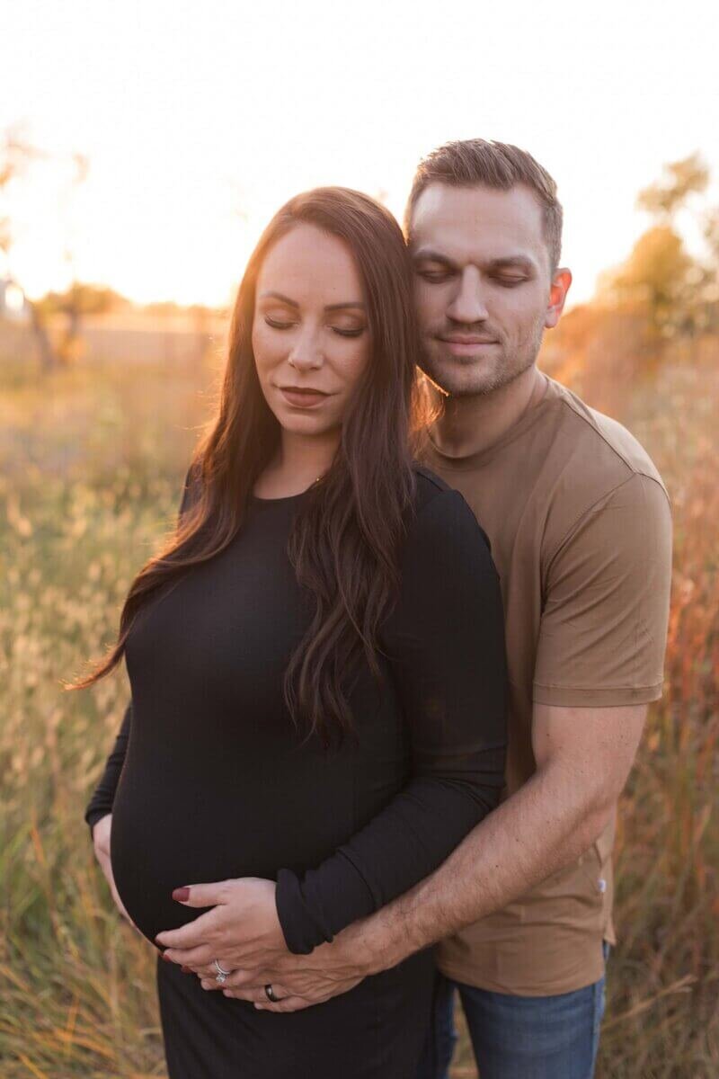 A couple stands in a field at sunset, with the man embracing the pregnant woman from behind as they both smile softly with closed eyes.