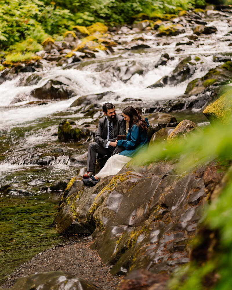 A couple in their wedding attire sits on the bank of a gushing river. they read their wedding vows during their olympic national park elopement