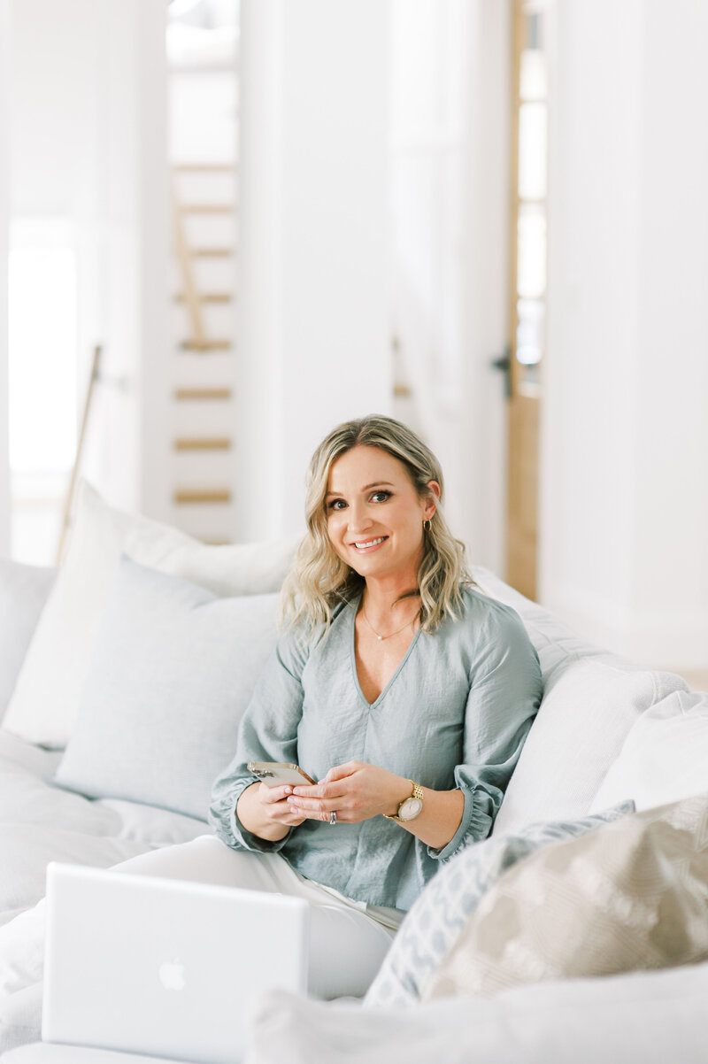 Jen Sottosanti, best London, Ontario interior designer and decorator sits on a couch in her living room with natural hues. She is holding her phone and smiling at the camera.