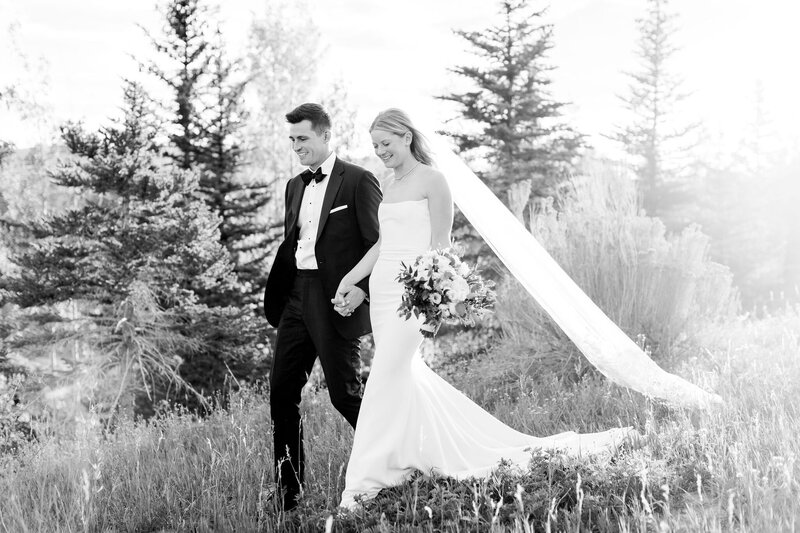 Black and white image of a bride and groom walking hand in hand through a field, as the brides vail trails behind and catches the golden hour light