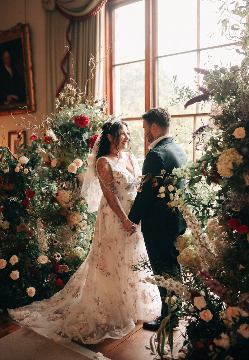 A bride poses and smiles surrounded by lots of greenery and flowers