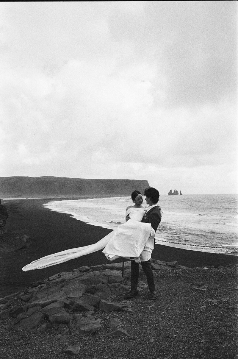 Groom holding bride in a flowing dress on a rocky cliffside overlooking a black sand beach, captured by a Spokane wedding photographer.