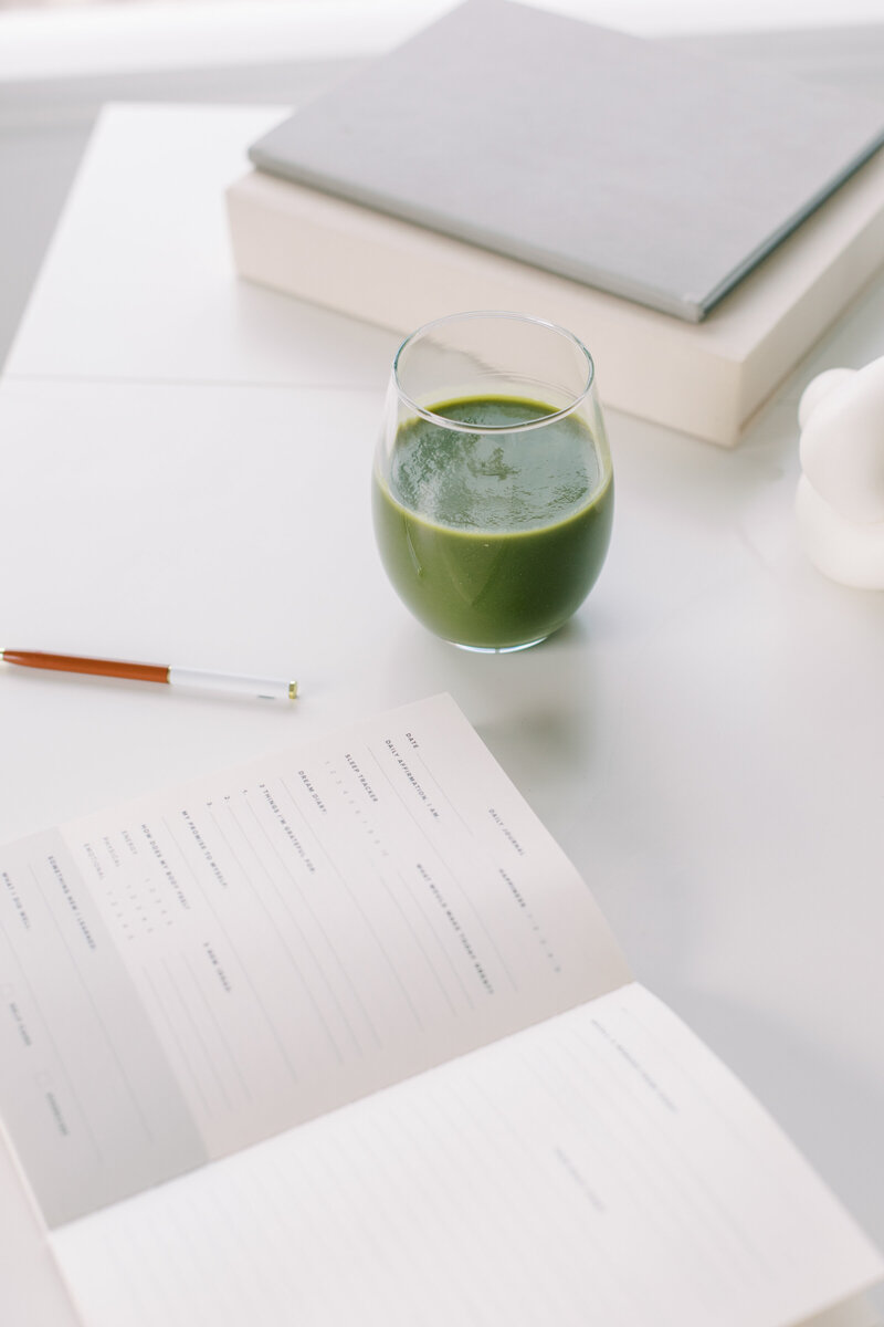 Glass of green juice beside an open wellness journal and a pen on a minimalist white desk.