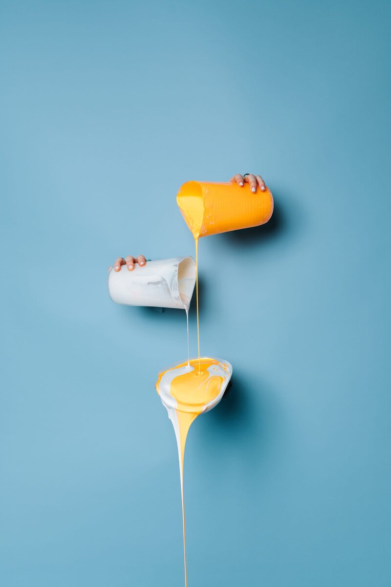 Aqua backdrop with 3 hands poking through pouring cups of white and yellow paint.