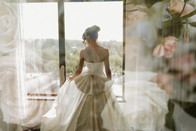 Bride in a satin gown standing by the window overlooking the city at Hyatt Regency Lisle in Illinois, captured by Rainstorm Photo & Video with a romantic, cinematic touch.