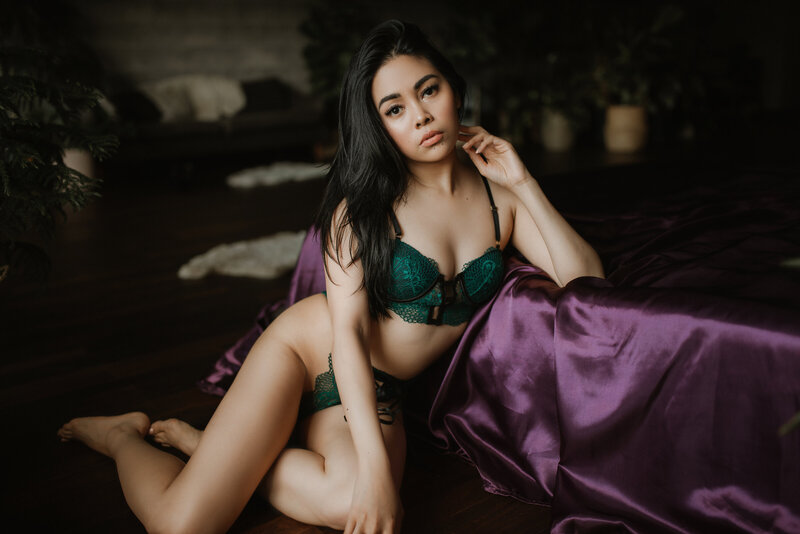 Woman in green lingerie poses on the floor beside a bed in the Vancouver, BC boudoir studio.