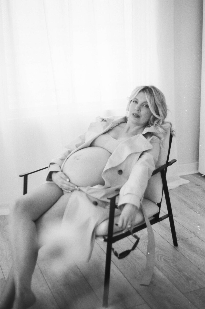 Black and white film photograph of a woman in a cream trench coat exposing her pregnant belly sitting in a modern chair