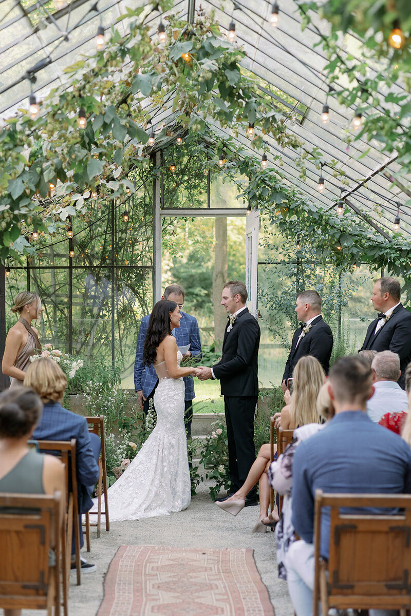 Bride and groom holding hands during their intimate ceremony inside the Glasshouse Community greenhouse filled with summer florals and natural light.