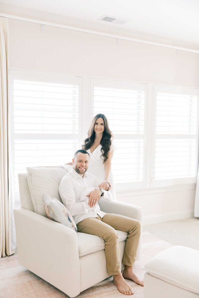 A father sits in a chair holding his sleeping newborn daughter smiling with mom leaning on the chair