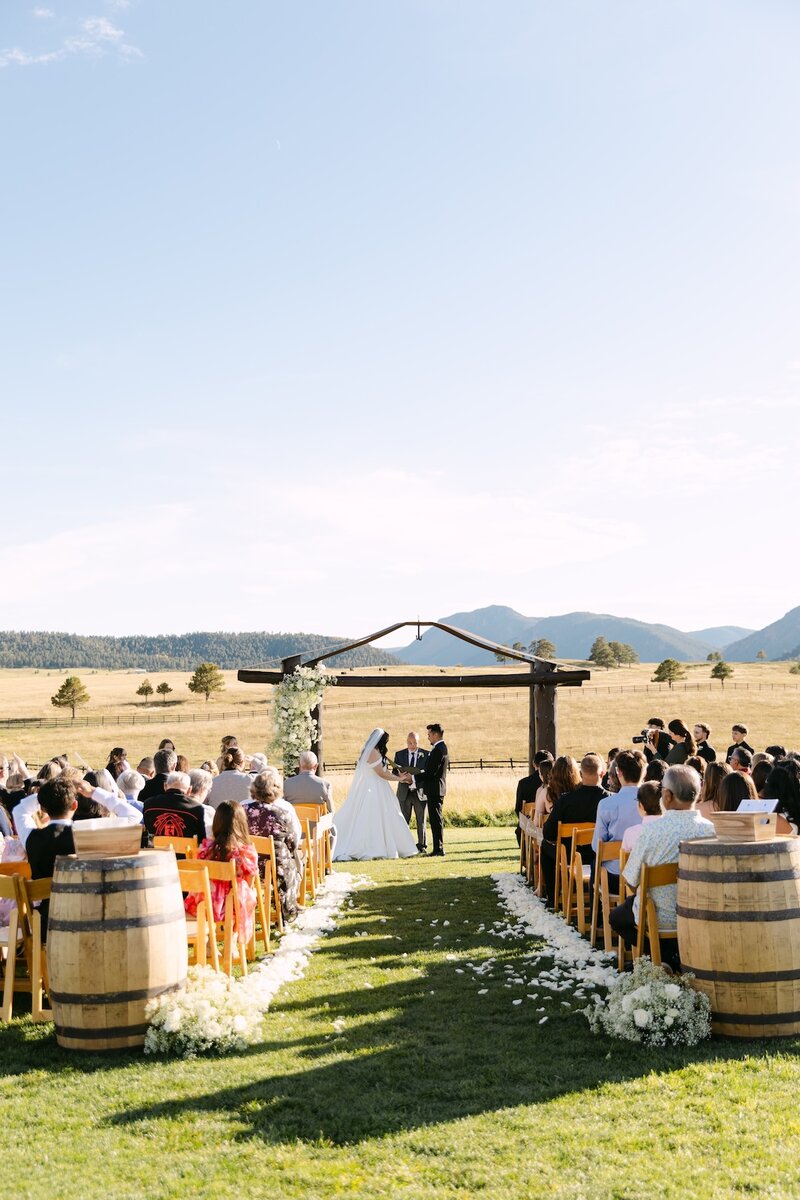 Outdoor Colorado wedding ceremony at Spruce Mountain Ranch with mountain views, wooden arch, and guests seated along floral-lined aisle — photographed by Colorado wedding photographer