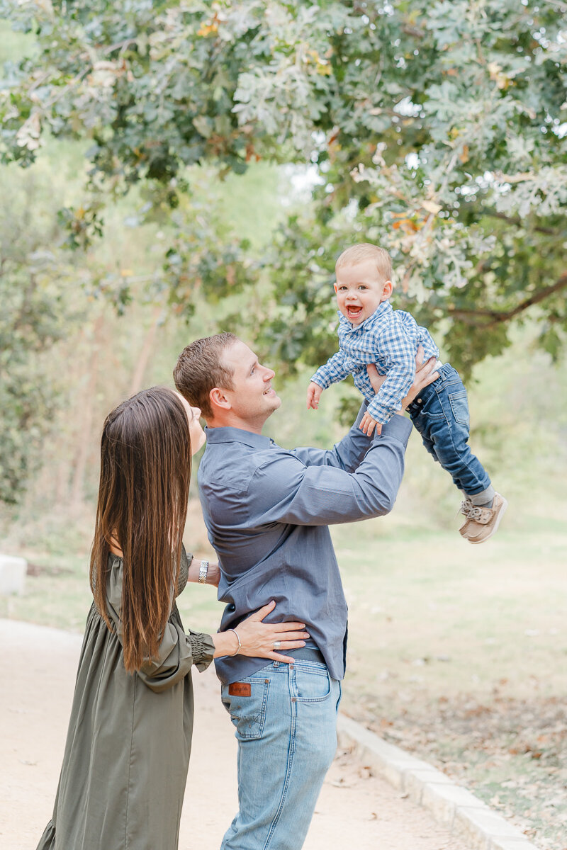 A mom and dad stand on a path at an Austin park. The dad holds their baby boy up in the air while his parents look up at him and smile.