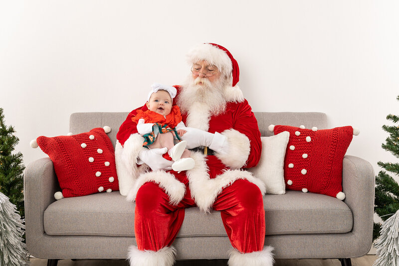 Baby sitting on Santas lap on a neutral couch with red pillows