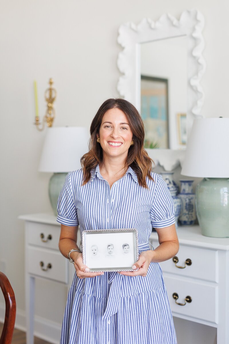 Woman in a striped dress holding a framed child’s portrait in a bright, styled room.