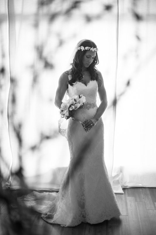 Black and white portrait of a bride in a strapless lace mermaid wedding dress holding a bouquet, standing by large windows at the White Room venue in historic downtown Saint Augustine