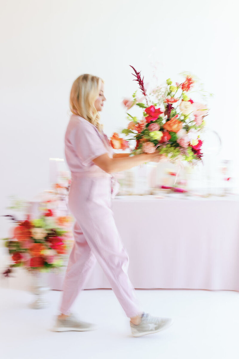 Motion blue photo of woman in light pink jumpsuit carrying a large colorful floral arrangement