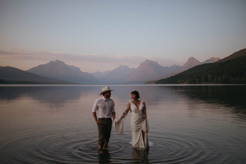 Bride and groom walk hand in hand through shallow water at Glacier National Park, surrounded by calm reflections and mountain peaks at sunset.