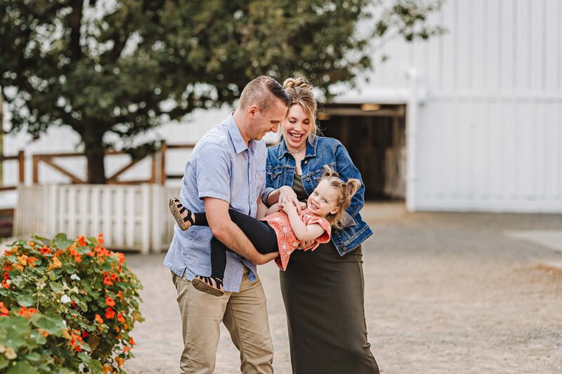 Parents lovingly holding their daughter at Kelsey Creek Farm in Bellevue, WA, with rustic farm scenery in the background.