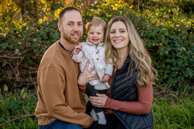 Mom and dad with their toddler smiling in a field of greenery at Pitt Lake in the fall.