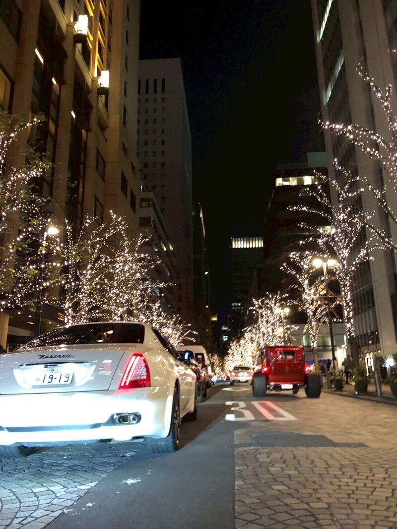 A white Maserati cruising down a sparkling, tree-lined street in Tokyo at night, with festive lights twinkling on both sides of the road.