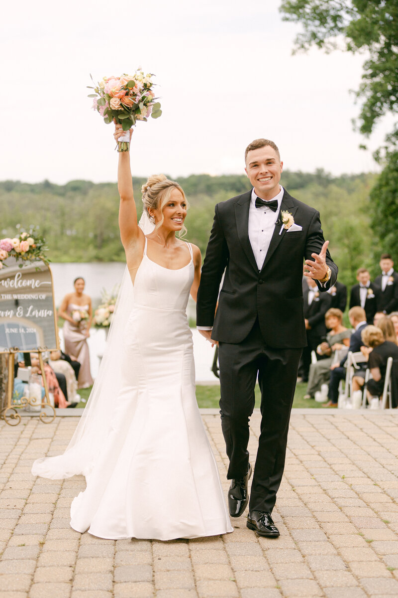 A bride in a white gown holds a bouquet triumphantly while walking beside a groom in a black tuxedo. They are outdoors, surrounded by guests and trees, with a lake in the background. A welcome sign is visible to the left.