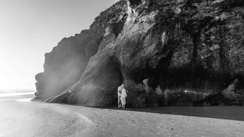 a black and white photo of a couple in their wedding attire as they embrace on a rocky coast during their adventure elopement. light leaks into the first third of the frame giving a dreamy quality to the image. 