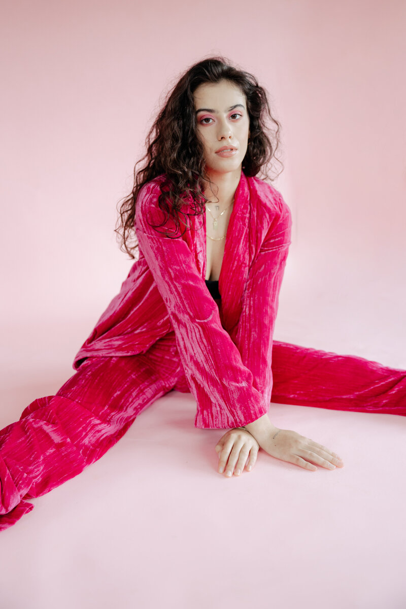 Portrait of a woman seated on a pink backdrop, wearing a vibrant pink textured suit, with a confident expression and natural curls.