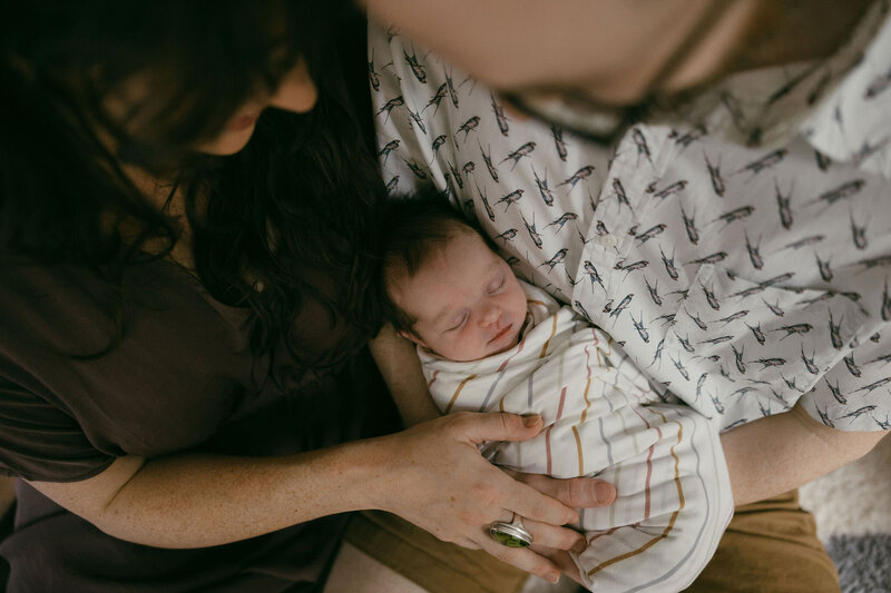 Beautiful, moody newborn photography session closeup of mom and dad overhead, looking down at sleeping baby boy swaddled in a striped blanket captured by Morgan Ashley Lynn Photography in Milwaukee, WI
