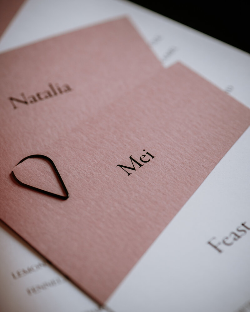 Pink wedding place card on textured paper, layered on top a white wedding menu and with a black clasp