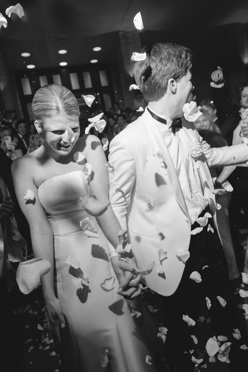black and white photo of couple walking through grand exit with flower petal toss, bride laughing down