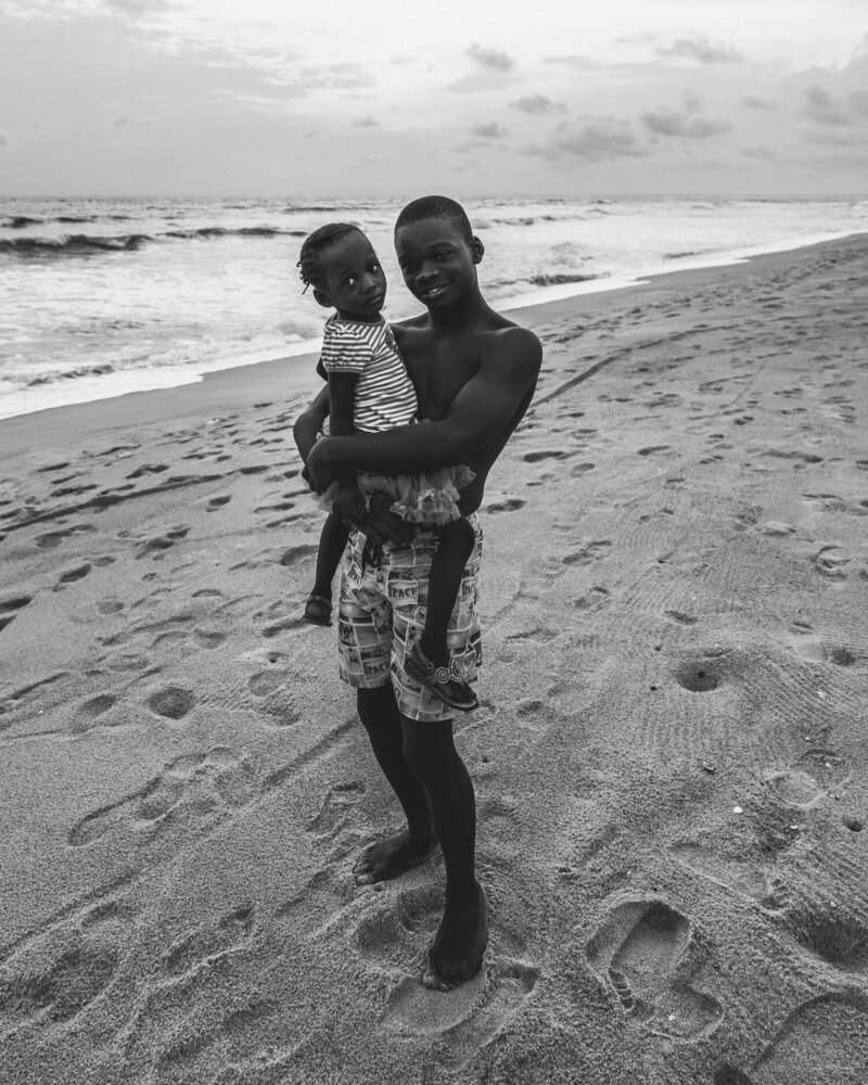 A Boy Holding A Young Child On A Beach In Liberia