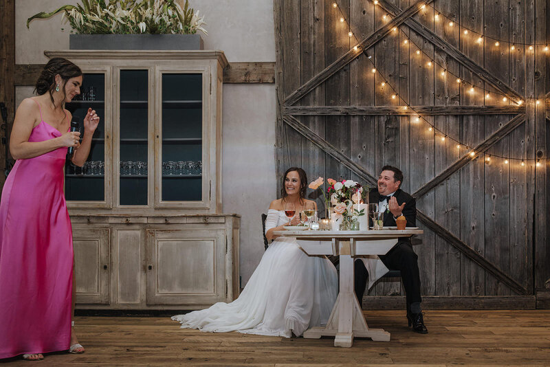 A newlywed couple sitting together as someone gives a speech