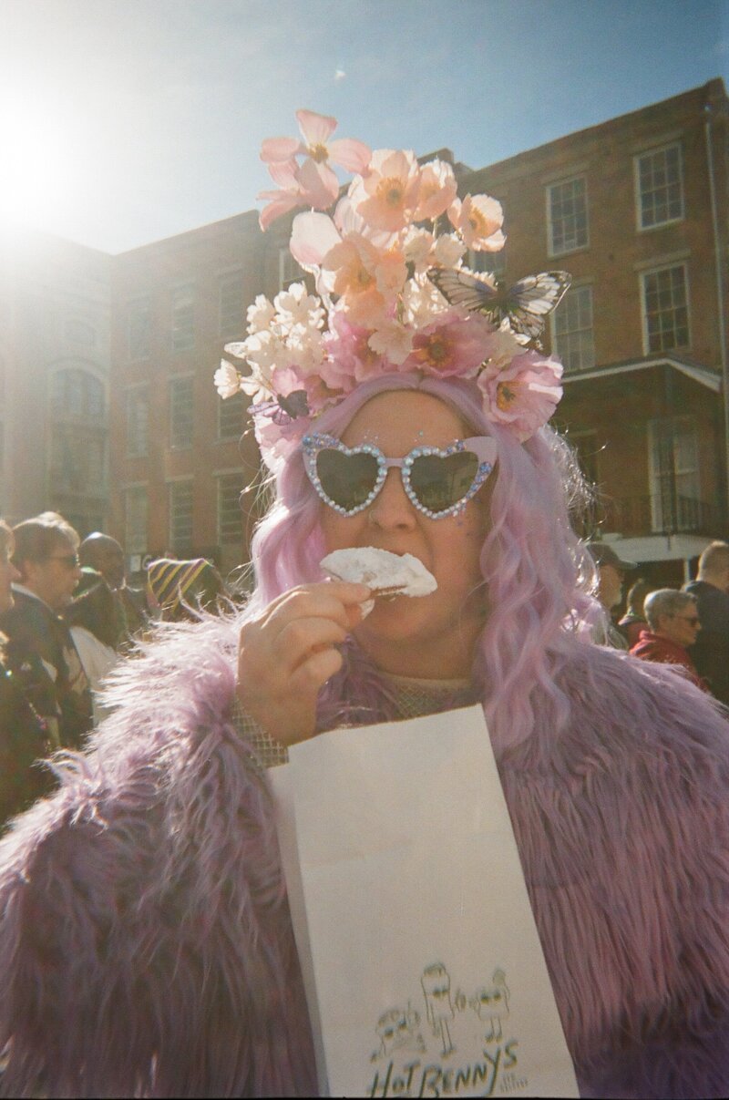 Olivia Yuen dressed in a lavender wig and flower crown, enjoying a beignet at a street festival.