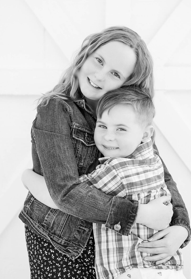 Brother and sister hugging each other and smiling by a small barn wall for North East, PA, family photography.