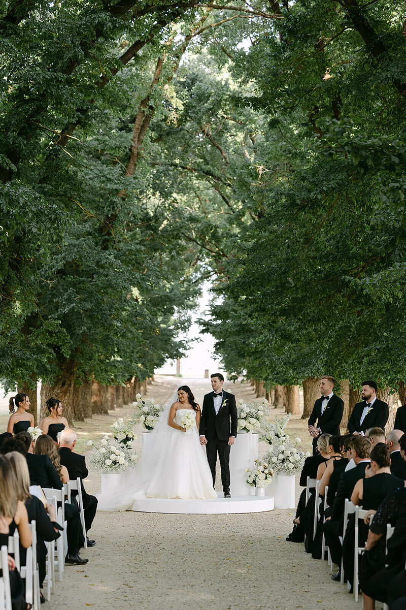 Bride and groom at ceremony on white stage under trees