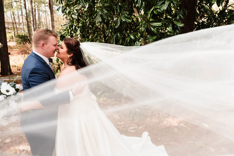 A bride and groom share a joyful embrace in an outdoor setting, surrounded by lush greenery. The bride’s veil flows dramatically in the wind, creating a dreamy, romantic moment. This stunning image captures the beauty and emotion of Alabama wedding photography.