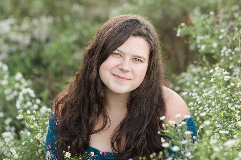 waynedale high school senior girl sitting in field of small white flowers with an off the shoulder green dress looking at camera smiling, photographed in holmes county ohio photographed by jamie lynette photography canton ohio senior photographer