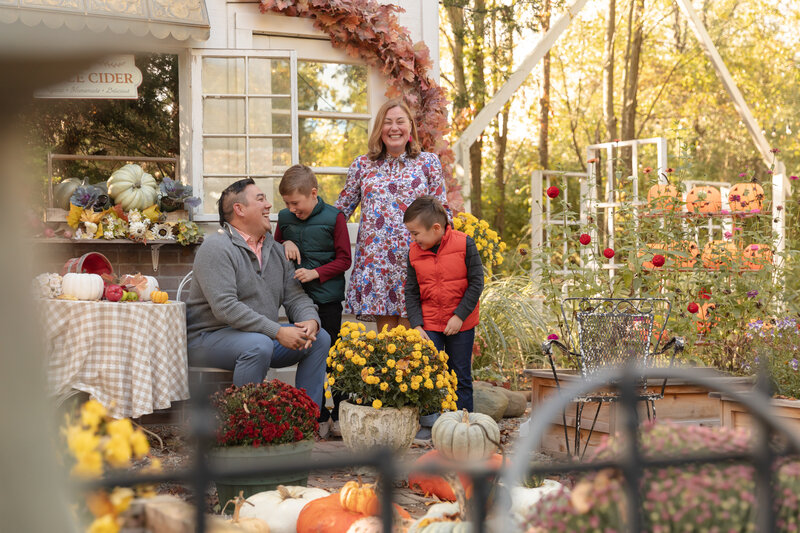 family of four gathers together at rustic wild arrow faux storefront and laughs surrounded by fall decor including leaves, apples, pumpkins and mums
