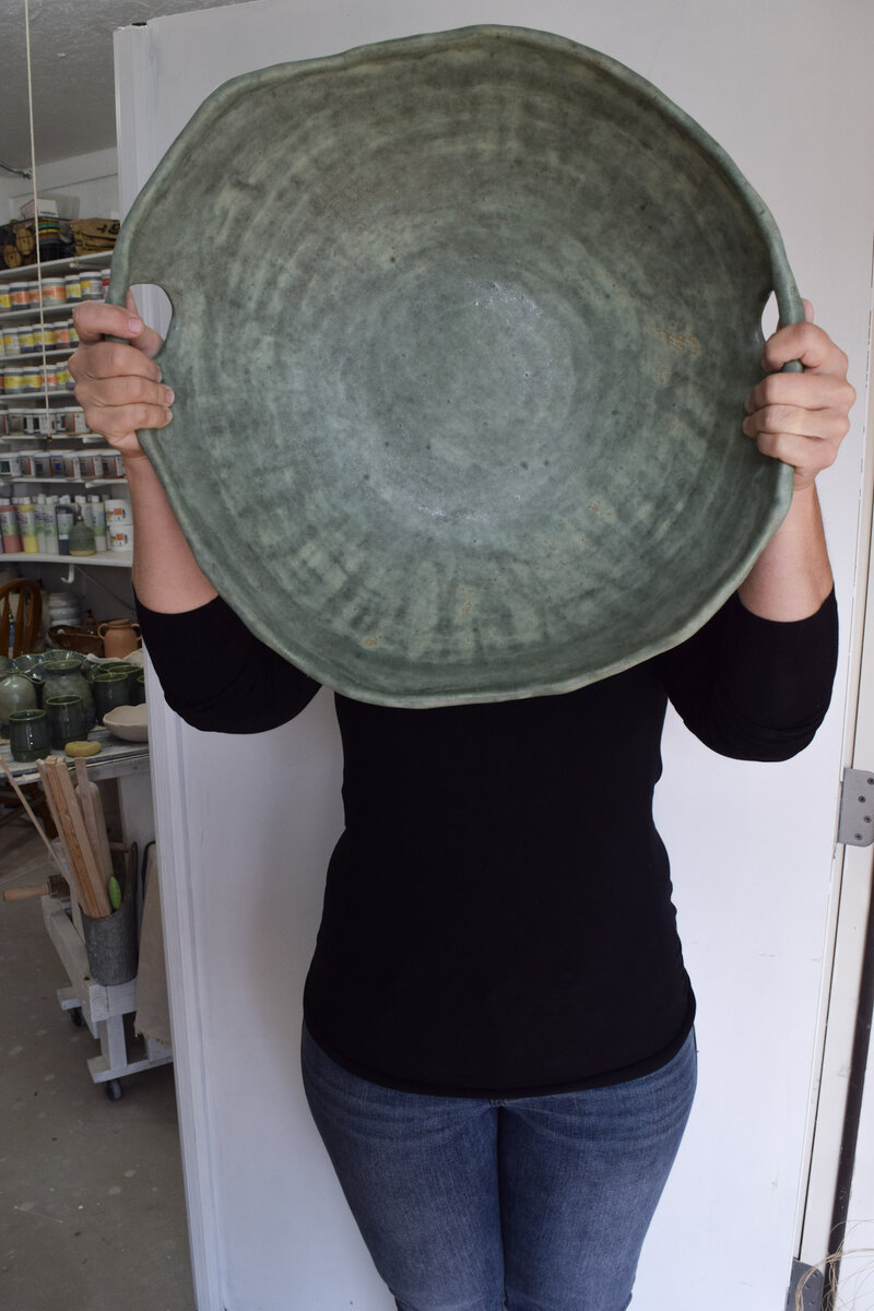 Jill holds a large green serving dish with handles in front of her face in her studio showing it's impressive scale