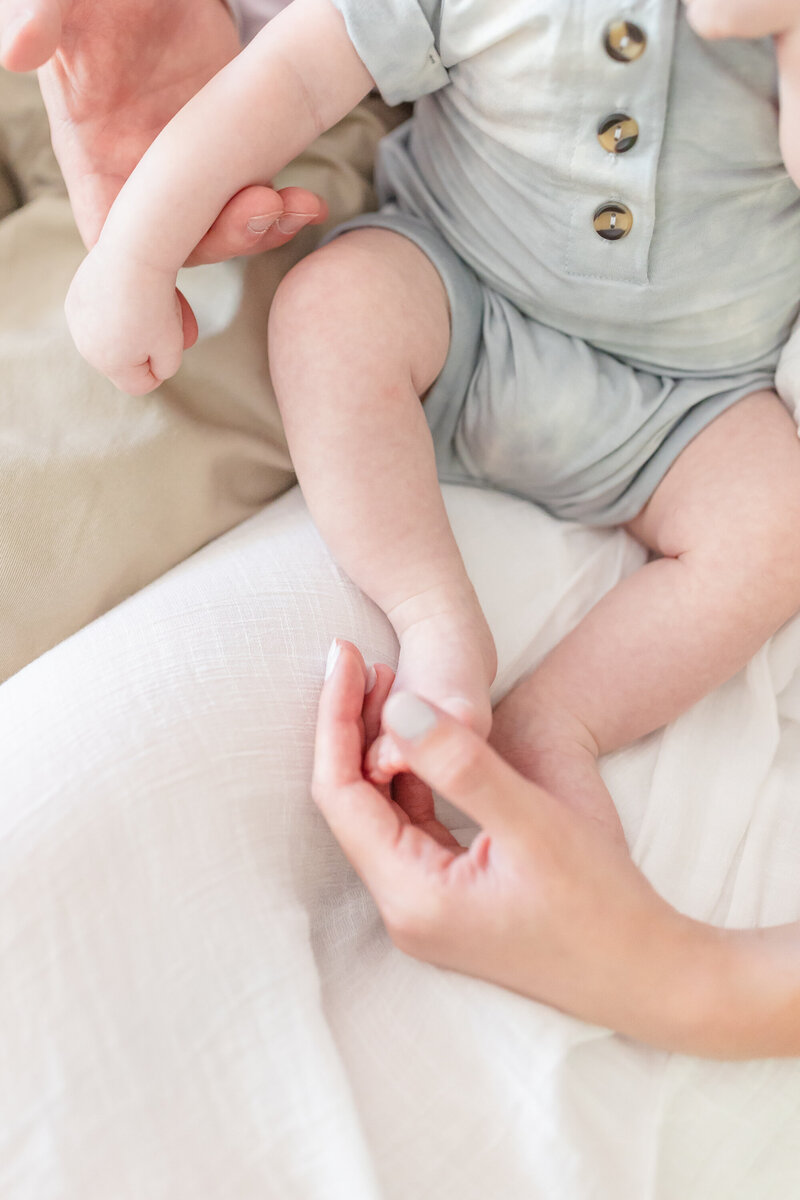  A delicate and intimate moment captured by a Connecticut newborn photographer, featuring a close-up of a baby's tiny hands and feet being gently held by their parents. The baby is dressed in a soft, neutral-toned outfit with button details, resting on a light fabric background, evoking warmth and tenderness.