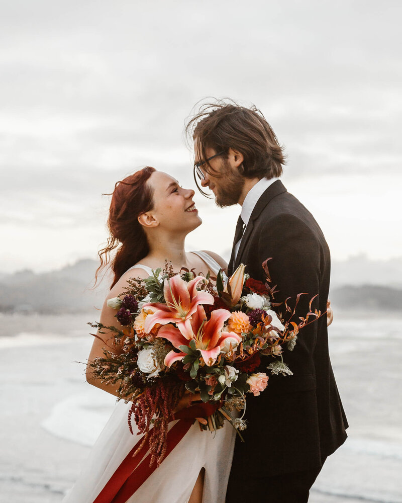 during their oregon coast elopement a bride holds her flowers and embraces her groom. They gaze lovingly at each other