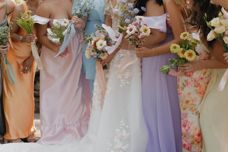 A wedding portrait of the couple with their entire wedding party. The couple is in the middle giving each other a kiss on the cheek and smiling. The wedding party is surrounding them looking excited and happy. The outfit colors are softer tones of pink, blue, orange, yellow, purple, and tan.