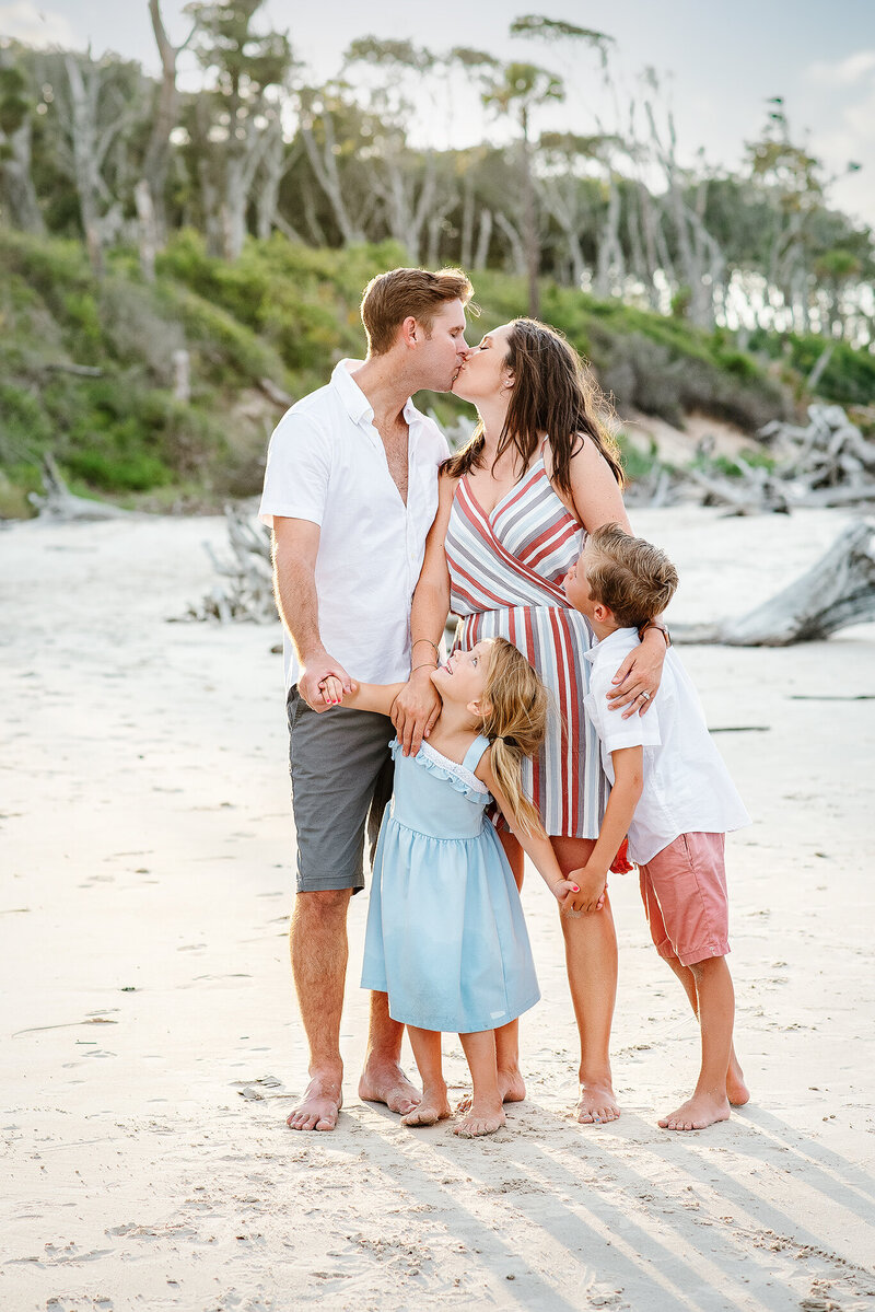 Brother and sister look up at parents kissing on beach at Talbot Island in Jacksonville, FL.