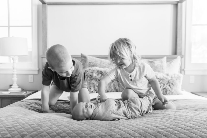Two young siblings smiling at their newborn baby brother wrapped in a blanket while sitting on a bed in a cozy bedroom