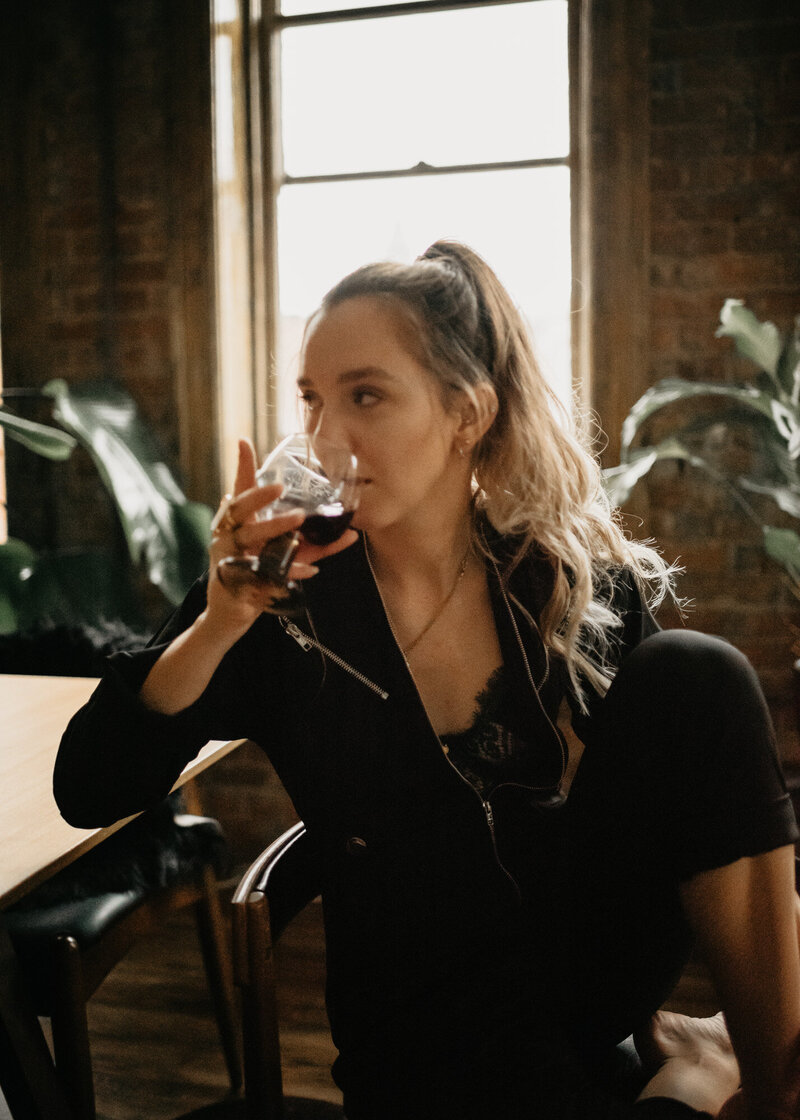 Showit websites designer, Clair Schwem, taking a break at her studio desk after working on a luxury branding project. She is sitting at a desk in front of a brick wall sipping red wine.