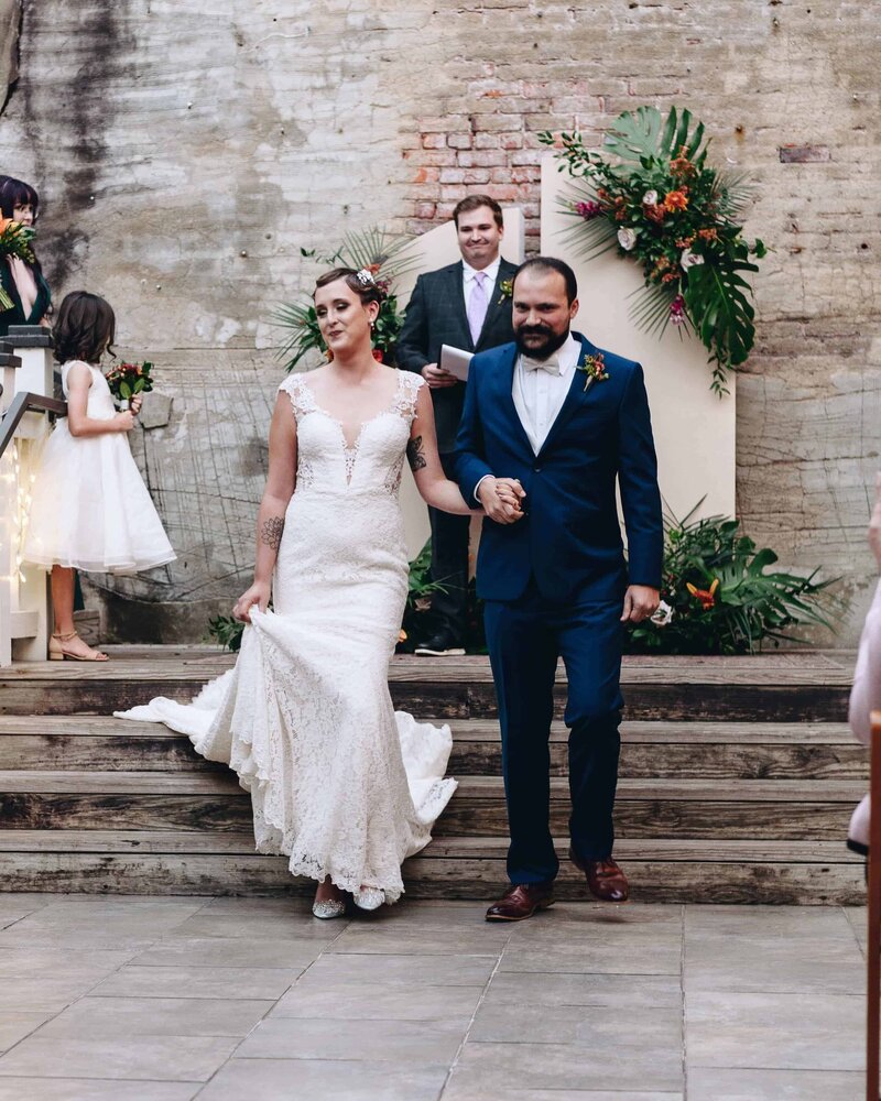 A bride in a white lace gown and a groom in a navy suit walk hand in hand down steps, smiling, after their wedding ceremony. A flower girl and officiant stand in the background near floral decorations.