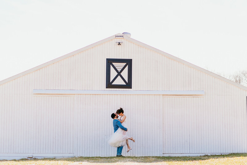 Newlywed couple sharing a dip kiss in front of a white barn during rustic Virginia elopement